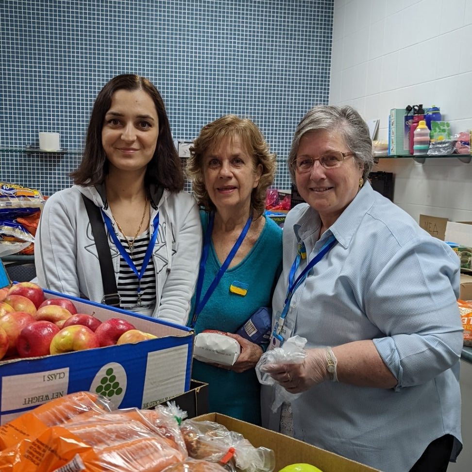 Harris Farm partners with Be Kind Sydney.  Image shows two volunteers each holding an apple standing in front of a delivery van with a stack of Harris Farm delivery boxes between them.